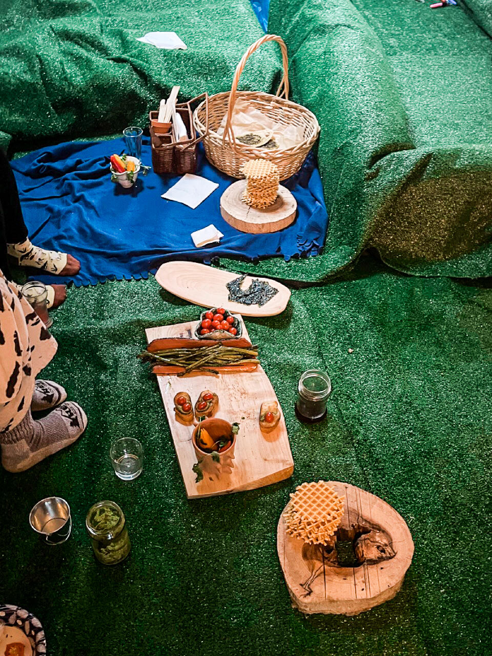 The green astroturf-covered floor of the apartment used for the indoor picnic. A blue blanket with a basket and a stack of waffle cookies, and other wooden food platters.