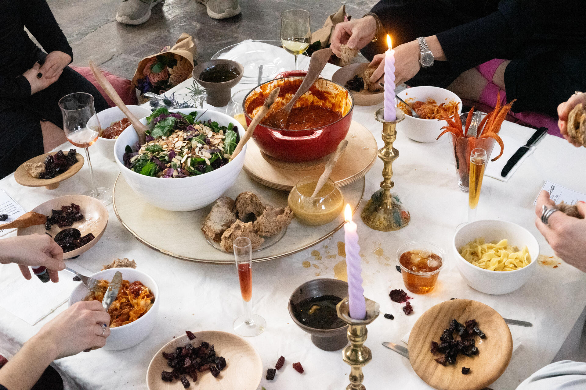 A close up detail photo of a low table with four people sitting around having a very colorful meal of salad, beets, chicken paprikash, purple candles, and with a wooden turntable in the middle. 