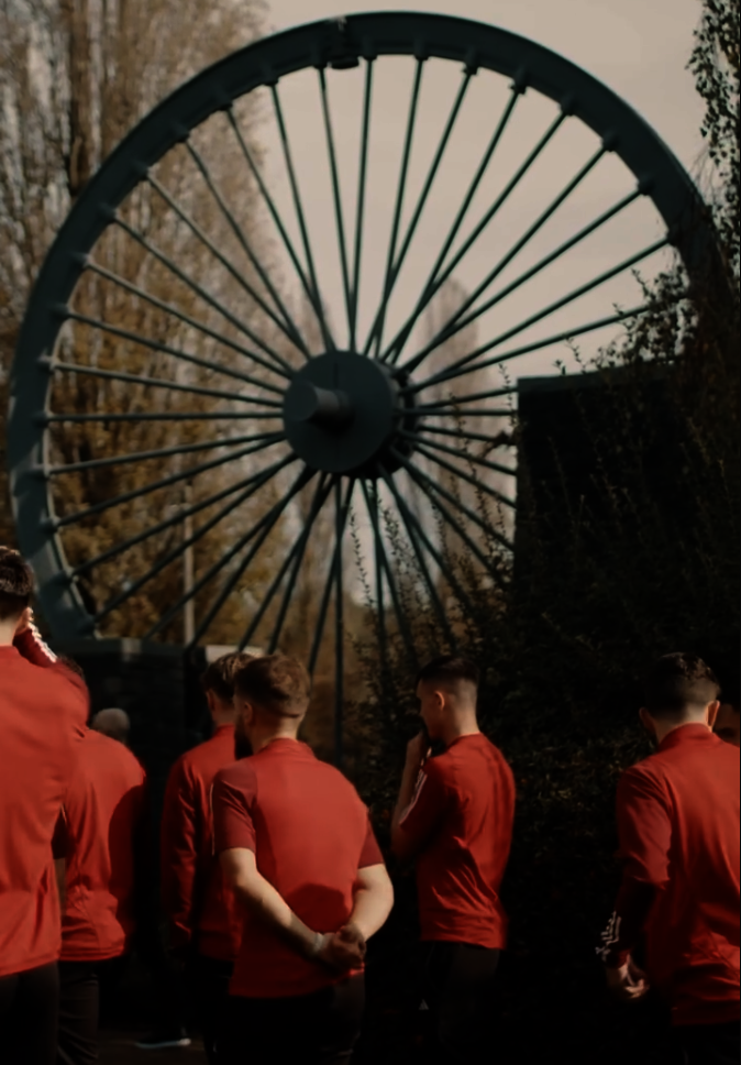 Welsh football players standing in front of a colliery wheel memorial to the Gresford mining disaster.