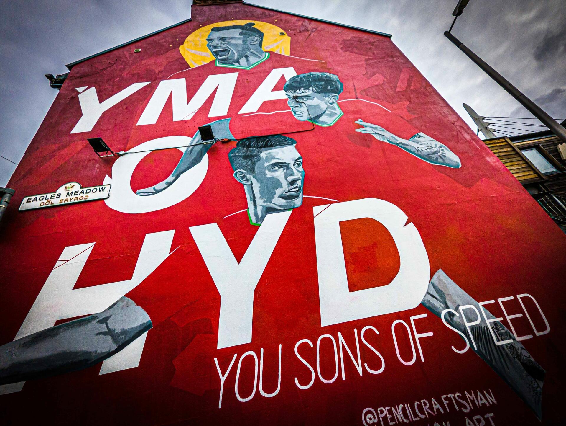 Mural of Welsh football players Gareth Bale, Neco Williams, and Harry Wilson on the side of a building with the words "Yma o Hyd You Sons of Speed."