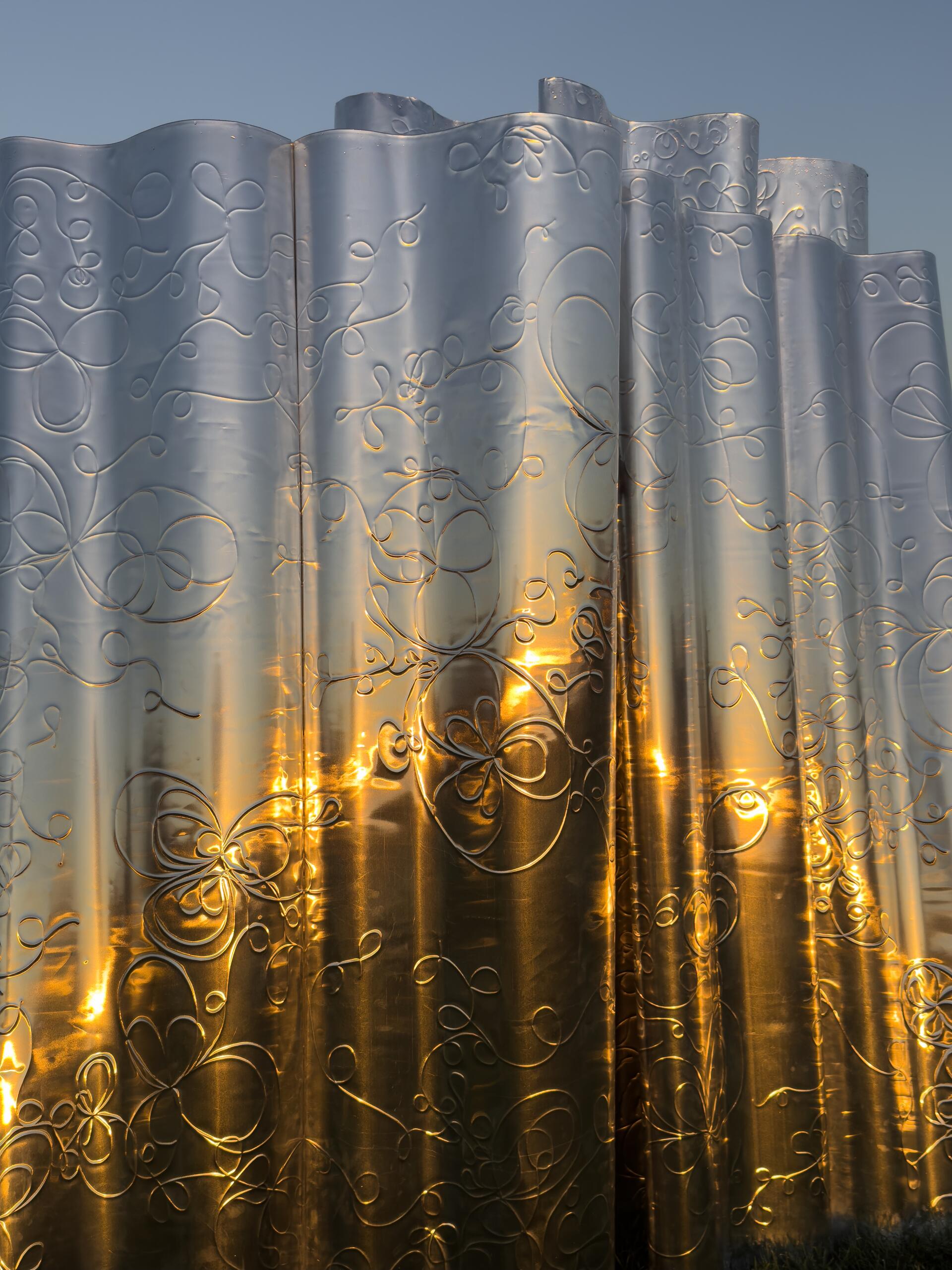 Detail of a metallic, wavy privacy screen with intricate floral patterns reflecting the golden hues of a sunrise. The surface creates a shimmering effect, capturing the light and adding texture to the smooth, undulating forms.