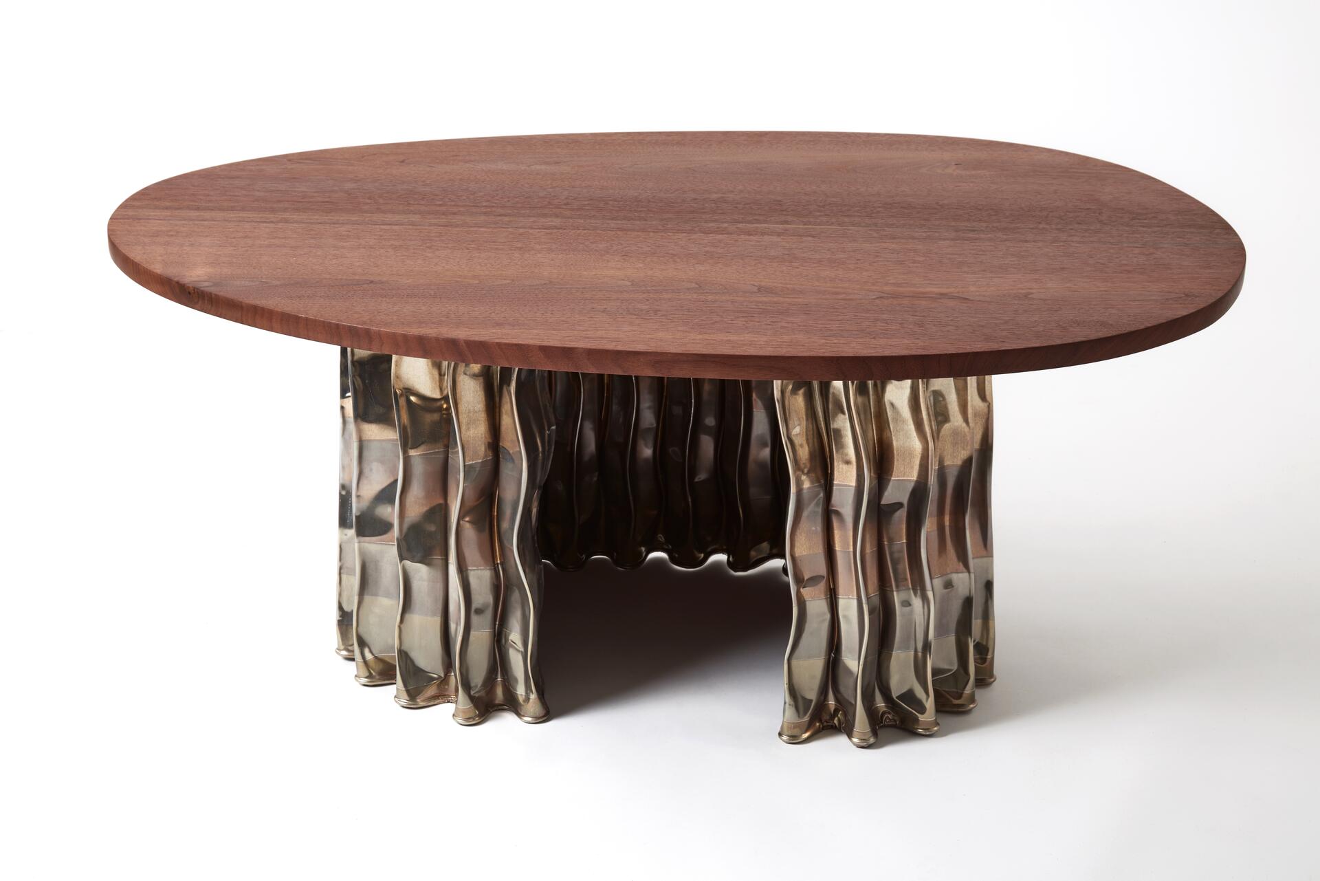 A round coffee table with a smooth walnut top and a unique, wavy metallic base. The base features a reflective surface with an elegant, flowing design, creating an artistic contrast with the wooden tabletop.