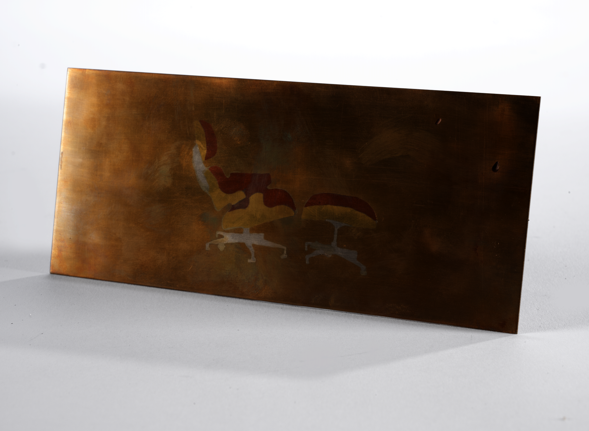 A thin rectangular sheet of bronze with an image in the center made of copper, brass, and silver depicting the side profile of the Eames Lounge Chair and Ottoman.