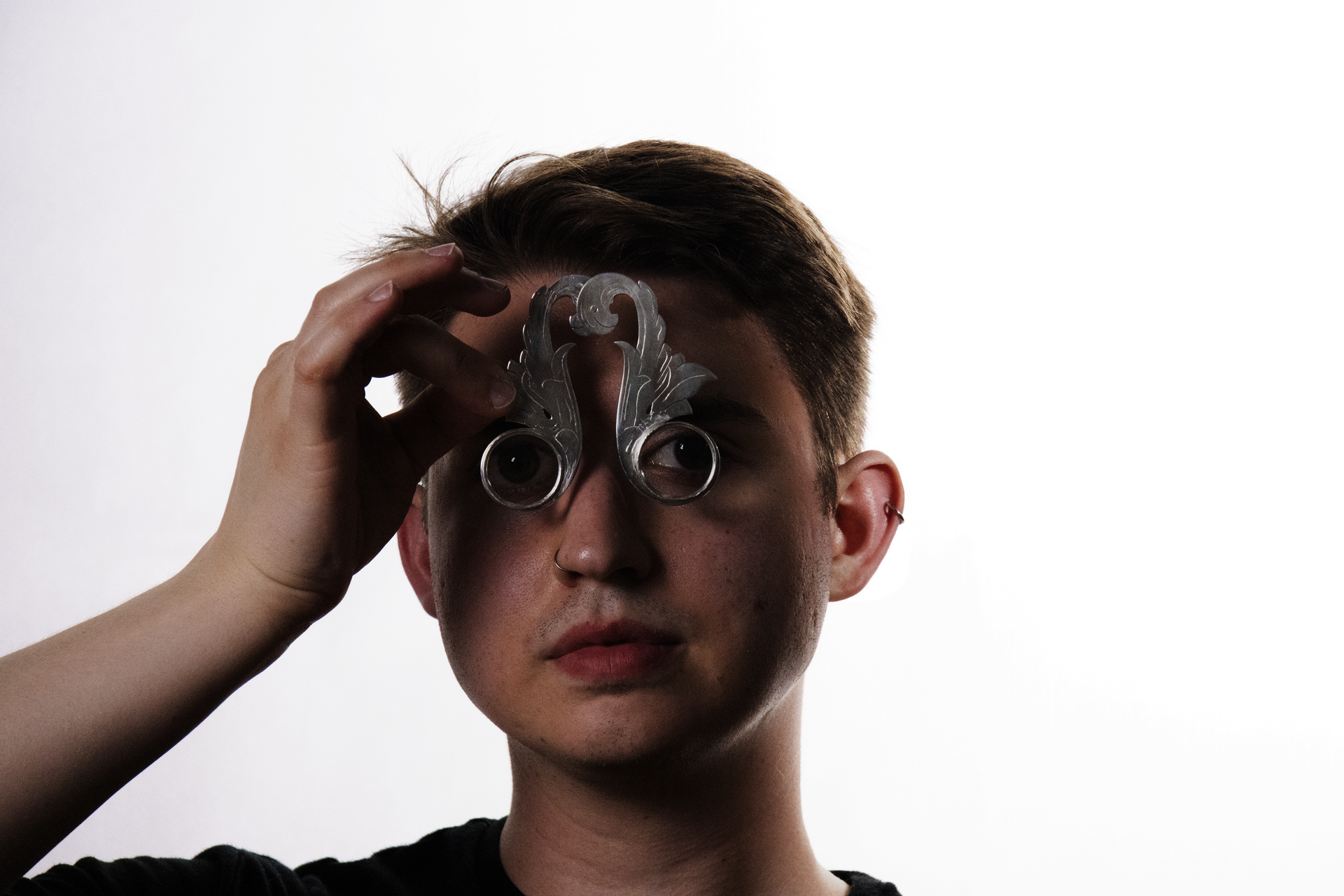 A man against a white background holds two magnifying glasses to his eyes held in silver frames that are cut and engraved to look like acanthus leaf illustrations and rivited together