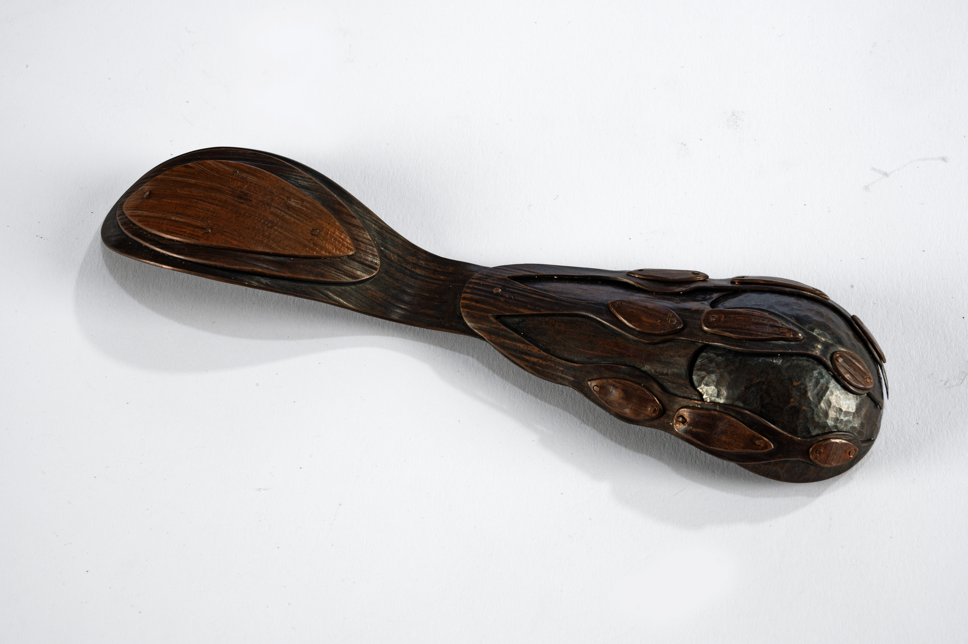 A spoon viewed from the underside made of copper with wood grain texture. Sheets of copper are cut, formed, stacked, and riveted together to look like a claw-and-ball chair foot