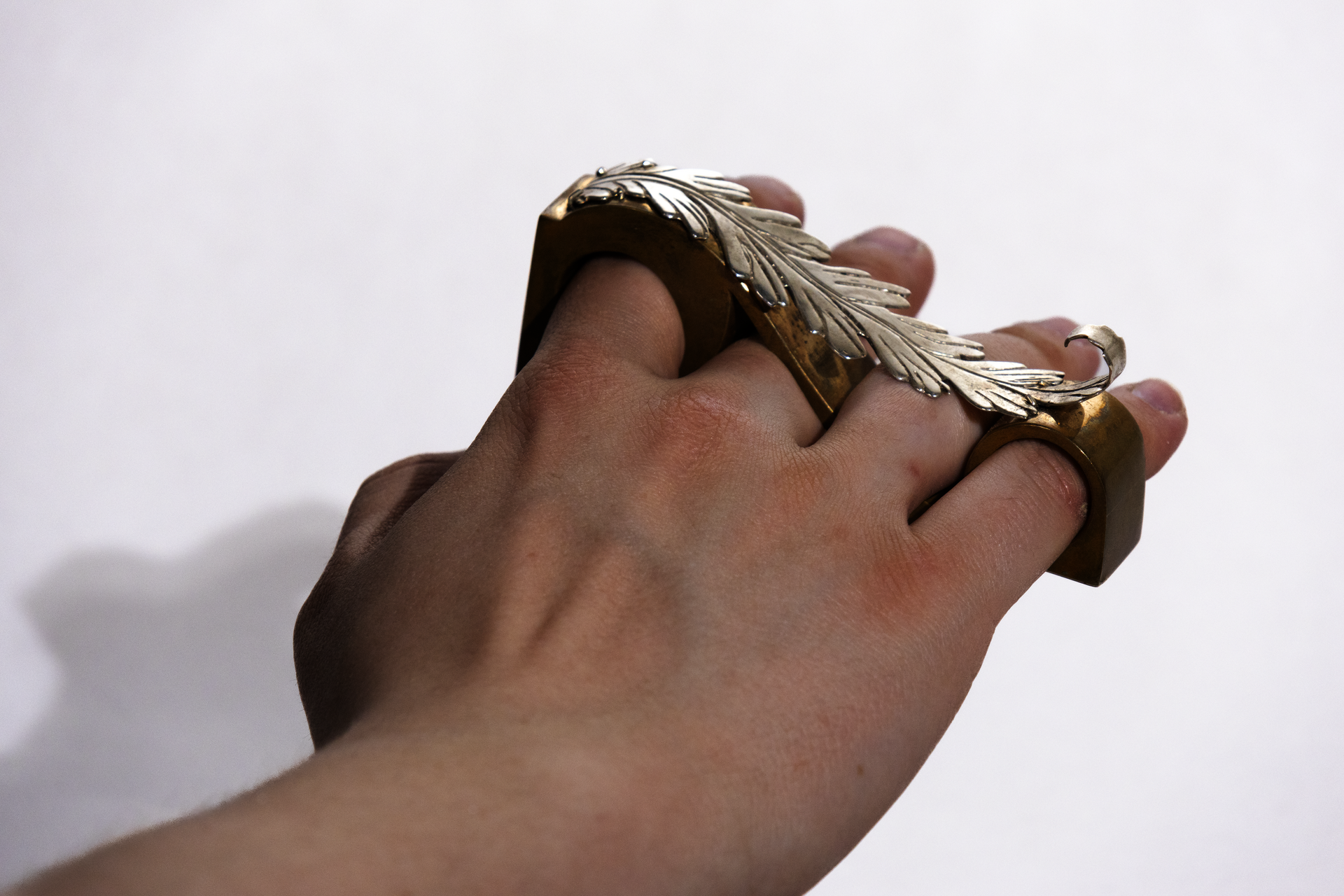 A hand reaching away from the viewer with a bronze ring that spans from the pointer to the pinky finger. The ring is capped with a curled silver acanthus leaf ornament.