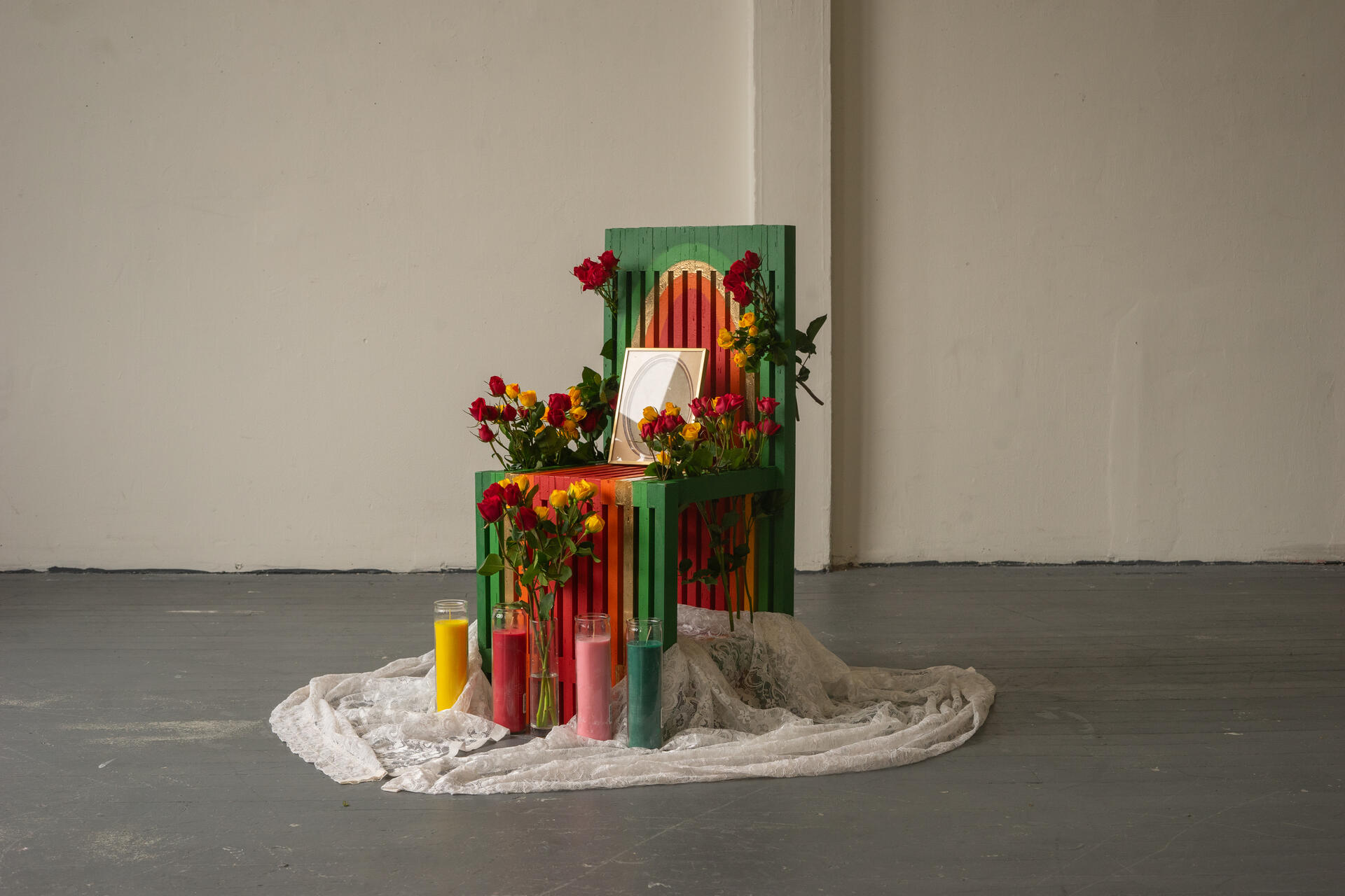 A slatted chair painted in a warm gradient sits in the middle of a stark room. Flowers are weaved between the slats, with a doilie, prayer candles, and flower vases sitting at its legs.