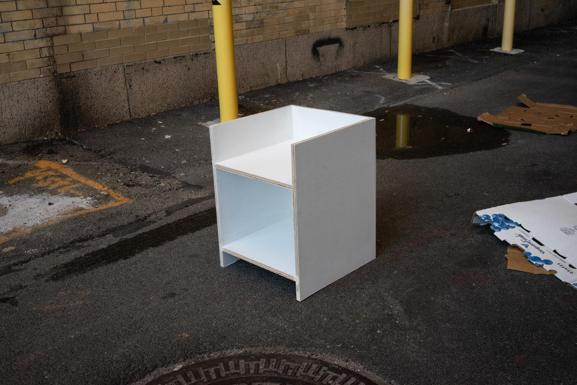A stark white chair in a rigid geomgetric form sits in a dark alleyway.