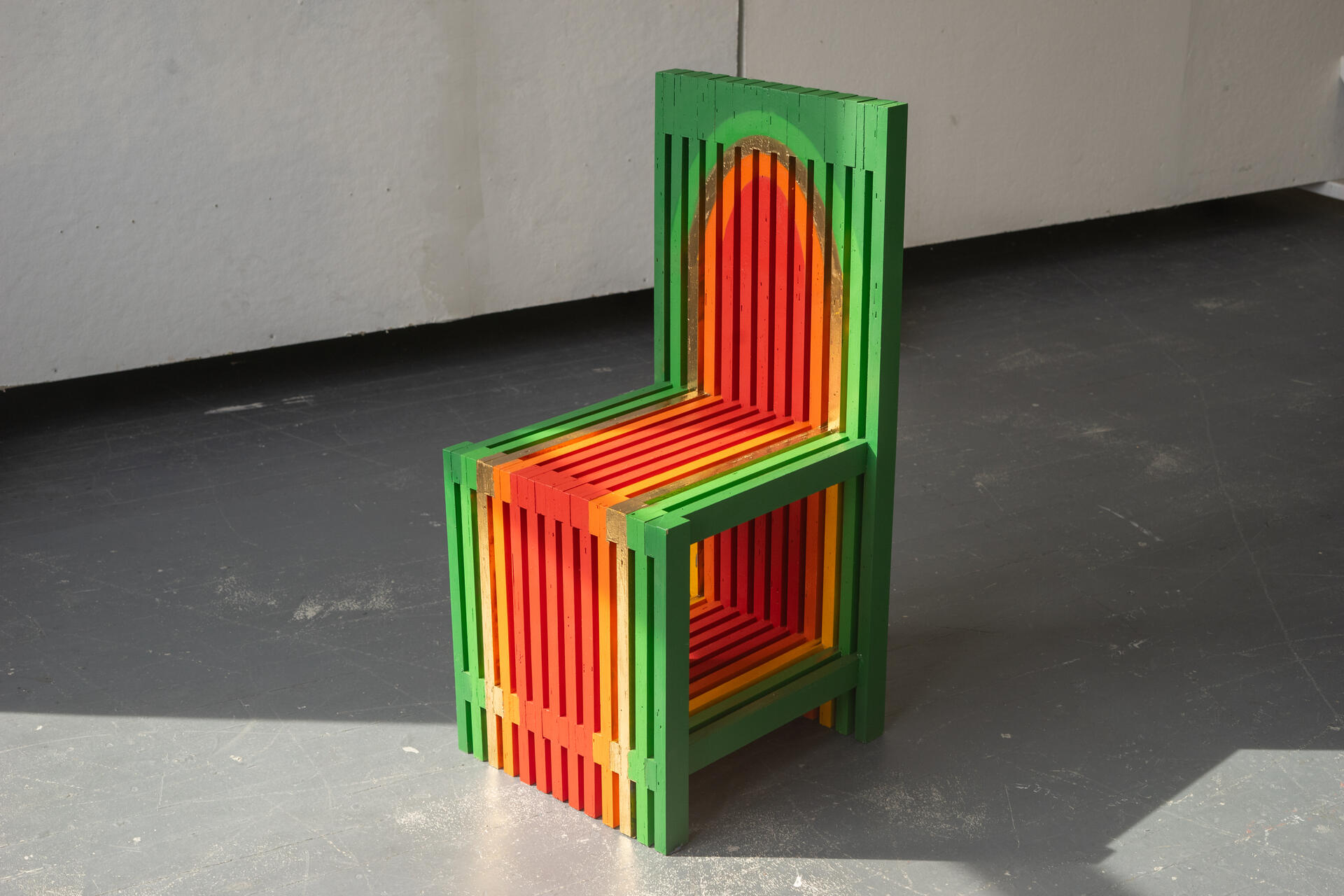 A colorful slatted chair painted in a colorful warm gradient sits in the middle of a stark room.