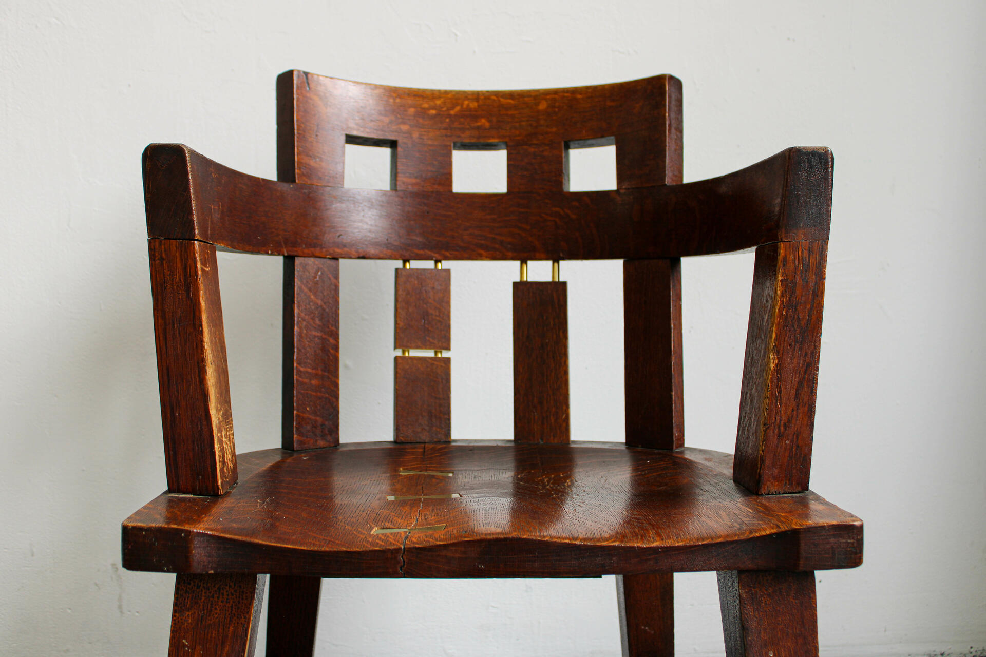 A weathered, dark brown oak chair is repaired with shiny brass metal. Brass bowties are embedded into seat where it cracked. Brass dowels show through several of the chair's legs and back dowels.