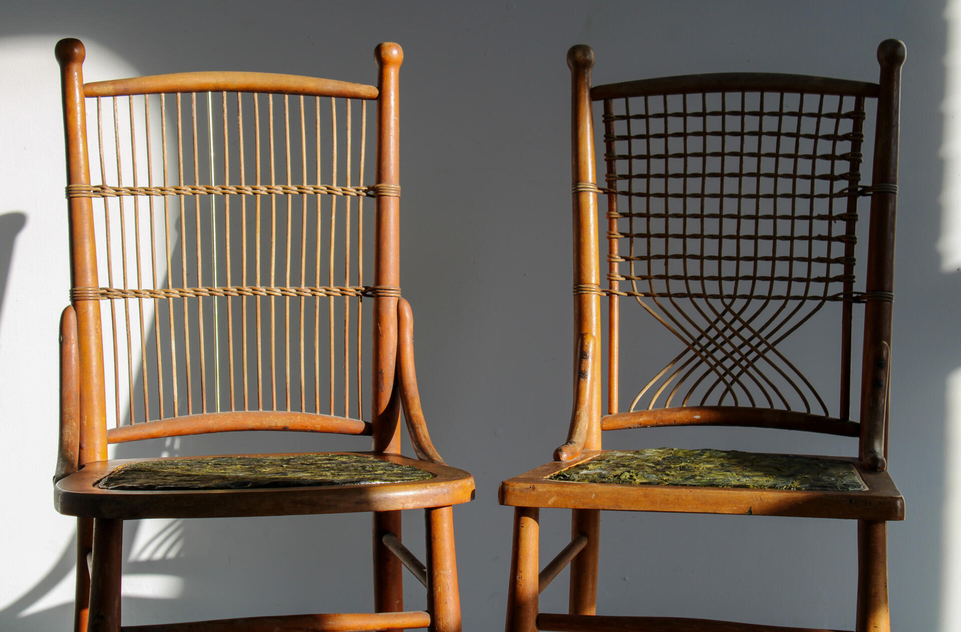 Two chairs (dark brown, ratan-style) sit side by side. Both have repaired seats made from woven pieces of kelp. The kelp reads various hues of green, from dark to light. Somewhat like a camoflauge pattern.