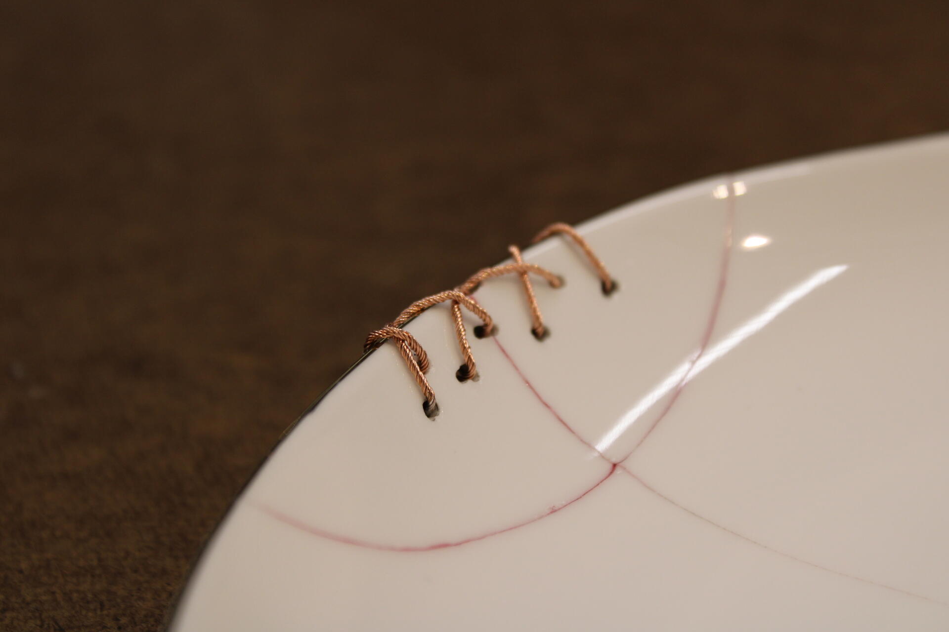 Close up image of the edge of a white plate which has been essentially sewn back together using tiny copper wires. The crack on the white plate is highlighted with a subtle red epoxy filling. 