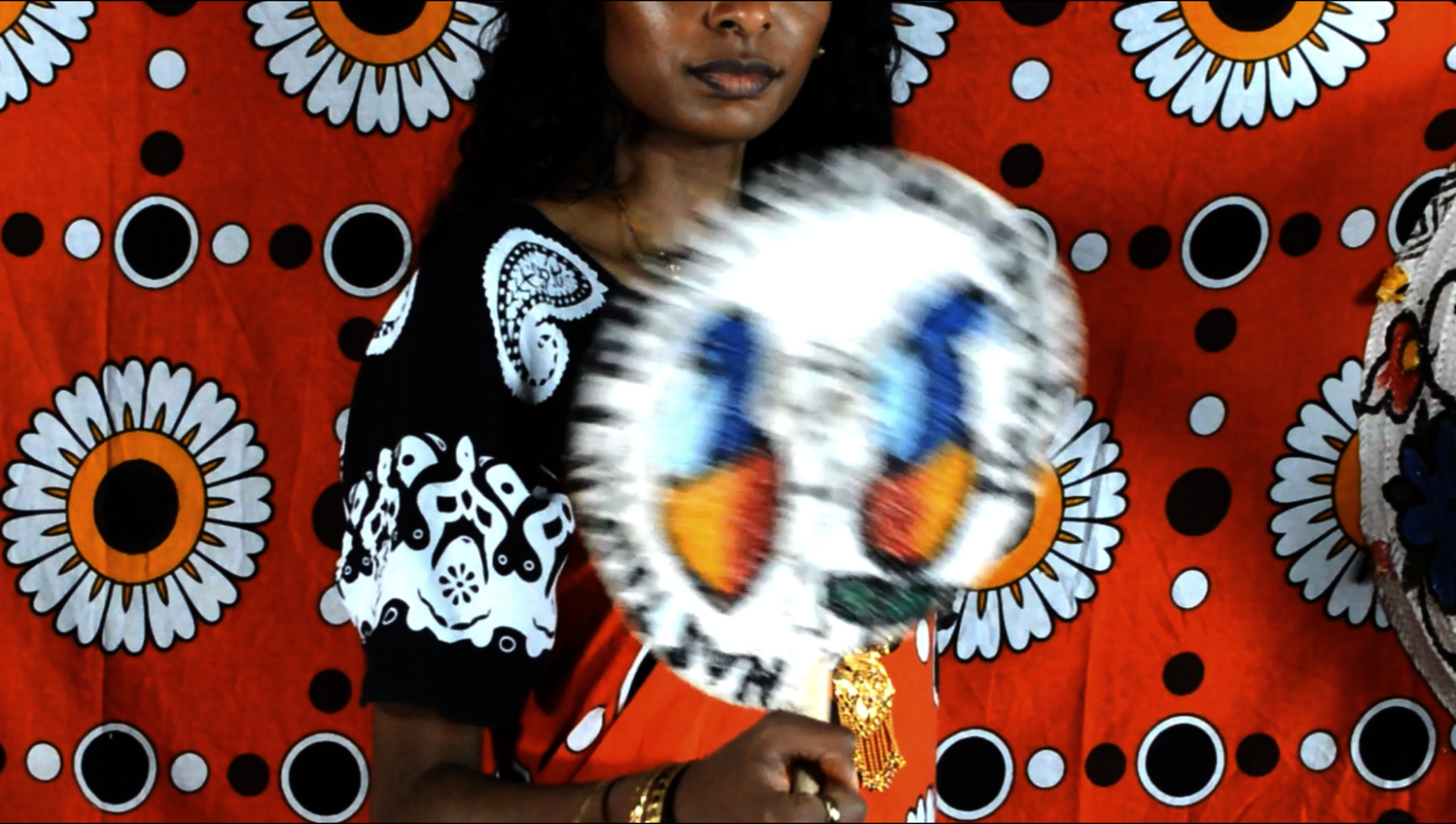 Screen shot of film from Siri Ya Mila of a woman holding a fan in front of a vibrant fabric.