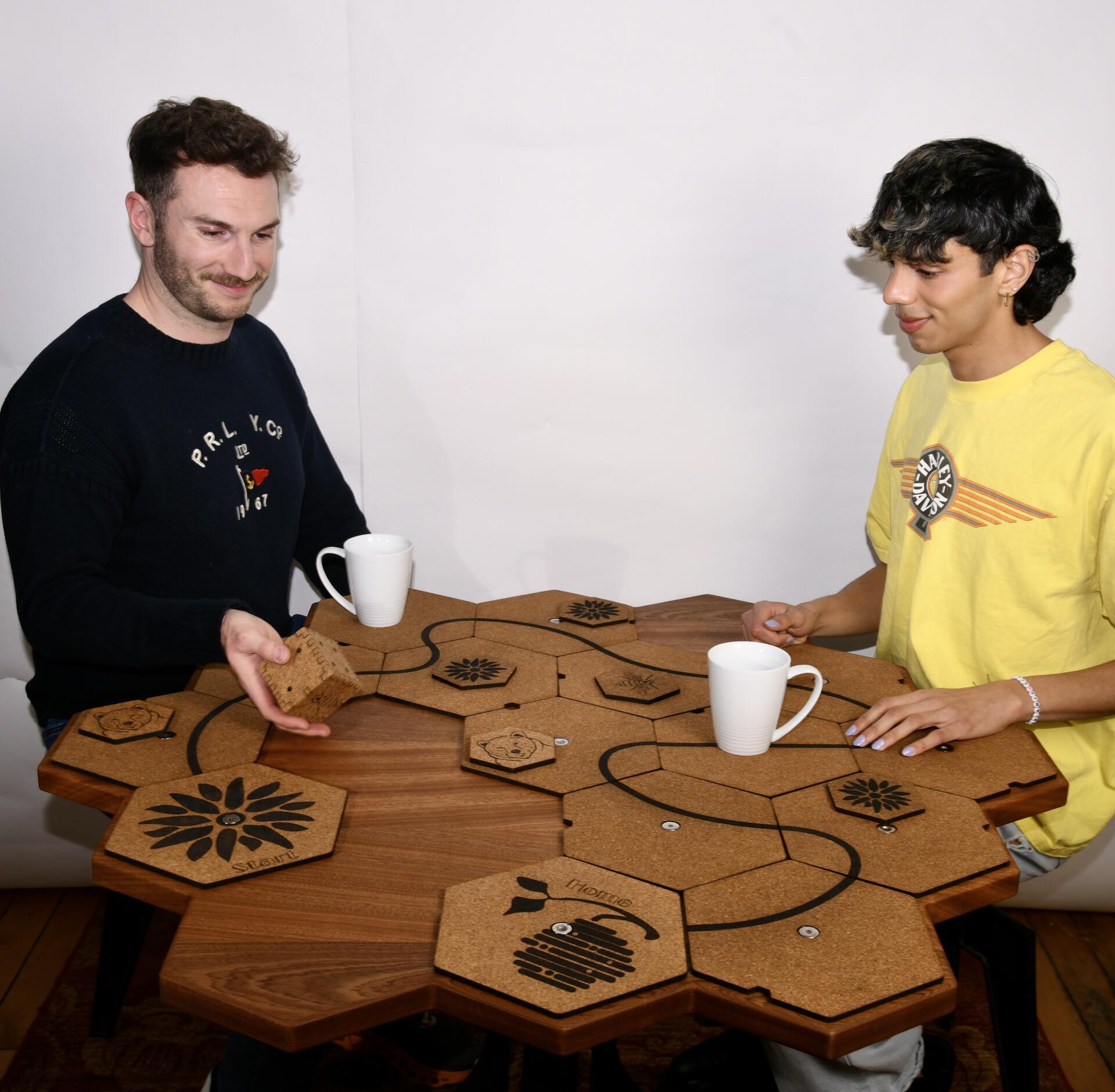 Two people, playing a game on the table, sapele table, honeycomb table, sitting together, one person moving cork tile, a white mug sitting on the table top