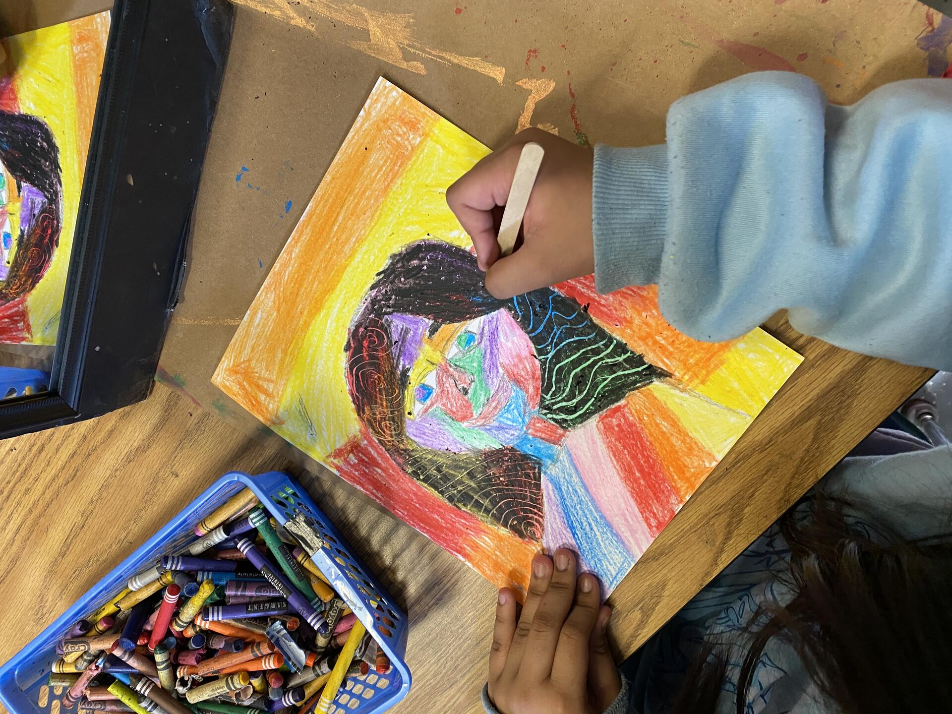 A student's hand holding a popsicles stick is scratching into a crayon-laden paper. The popsicle stick is creating patterns in the self portrait. A bin of crayons can be seen to the left of the image.