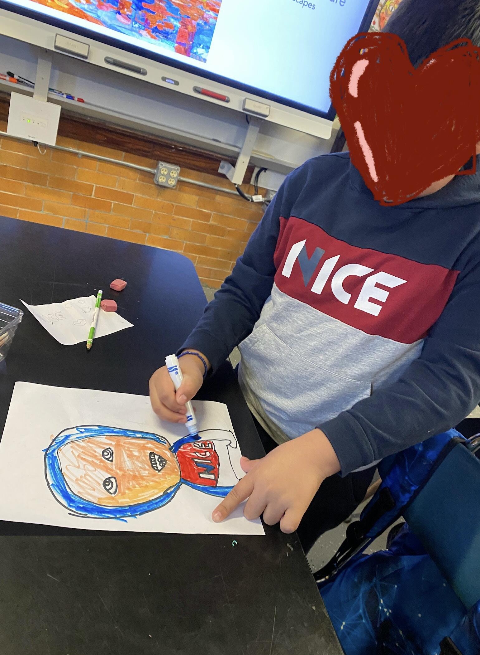 A student with a heart over their face to protect privacy is standing at a table and coloring with marker a portrait of themselves. The student is using blue, red, and a tan colored marker. 