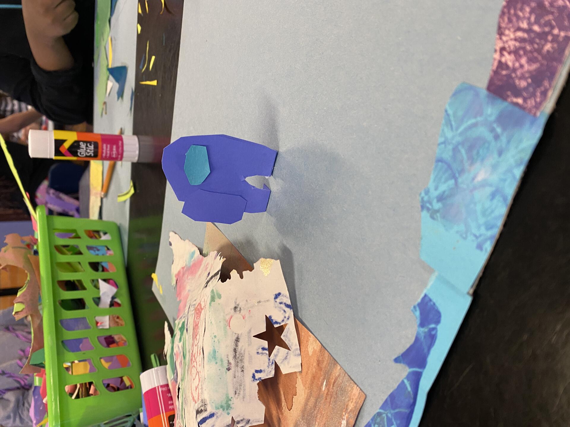 A paper collage cut out of a spaceman from the video game Among Us stands up in a 3D fashion on a student's paper. Seen in the background is a mess of papers and gluesticks.