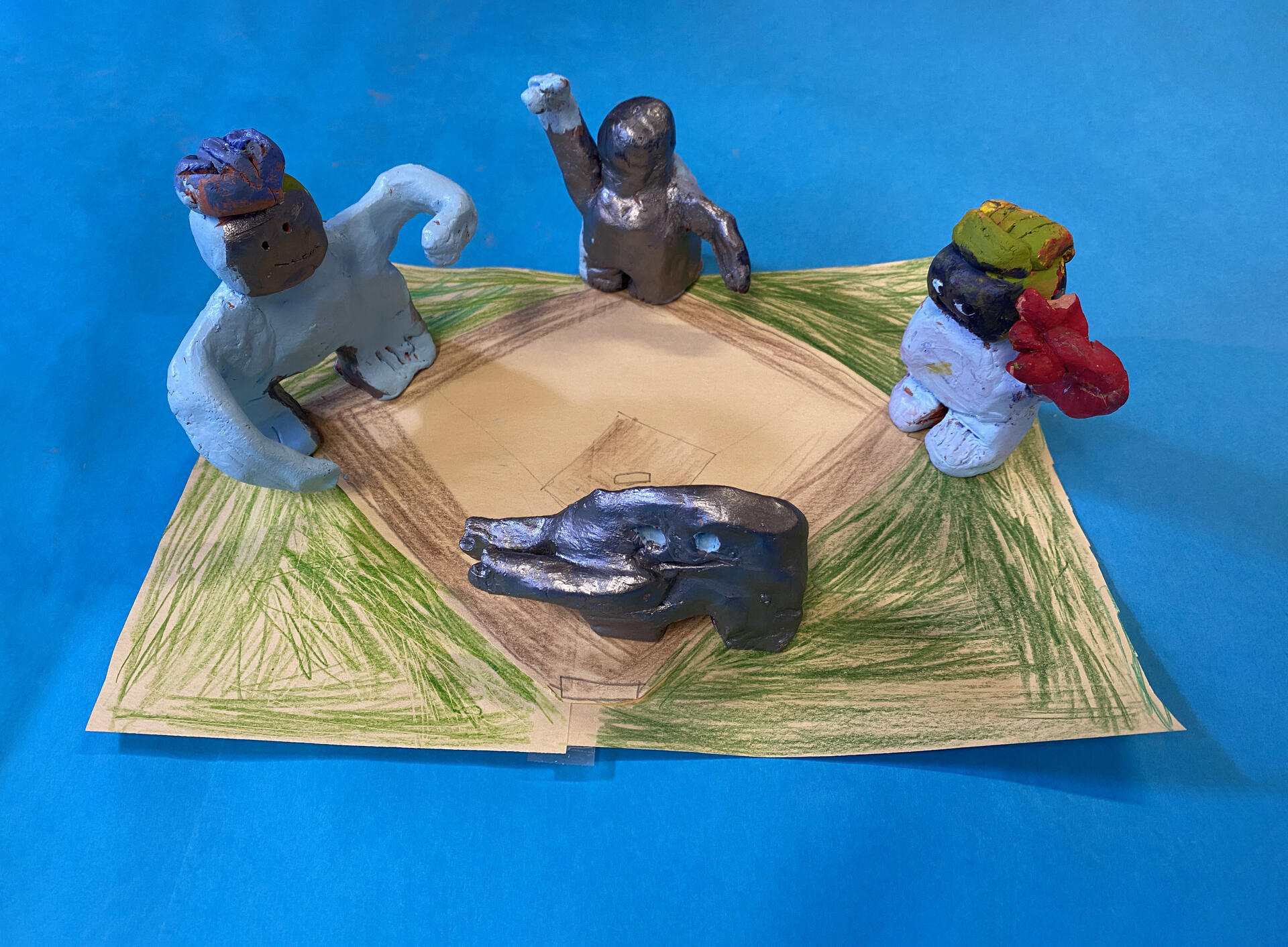 Four clay figurines stand on a hand drawn baseball diamond atop a bright blue backdrop. The figurines have been painted a combonation of silver, light blue, and red, and all have some version of outstretched arms.