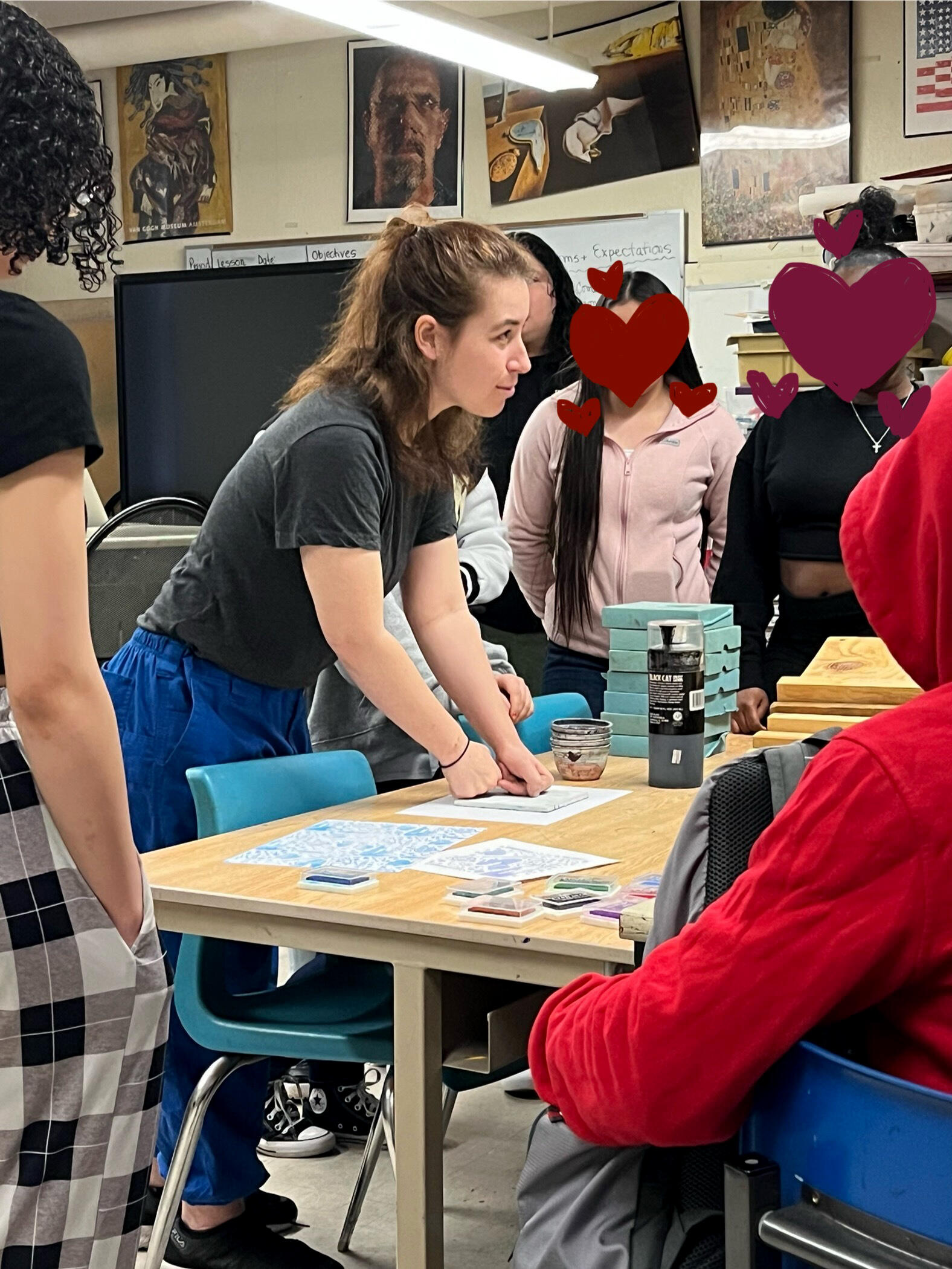 A woman in the center of the image is surrounded by a group of high-schoolers. The woman is pushing down on a sandwich of thin easy-cut linoleum block and a piece of paper onto the table.