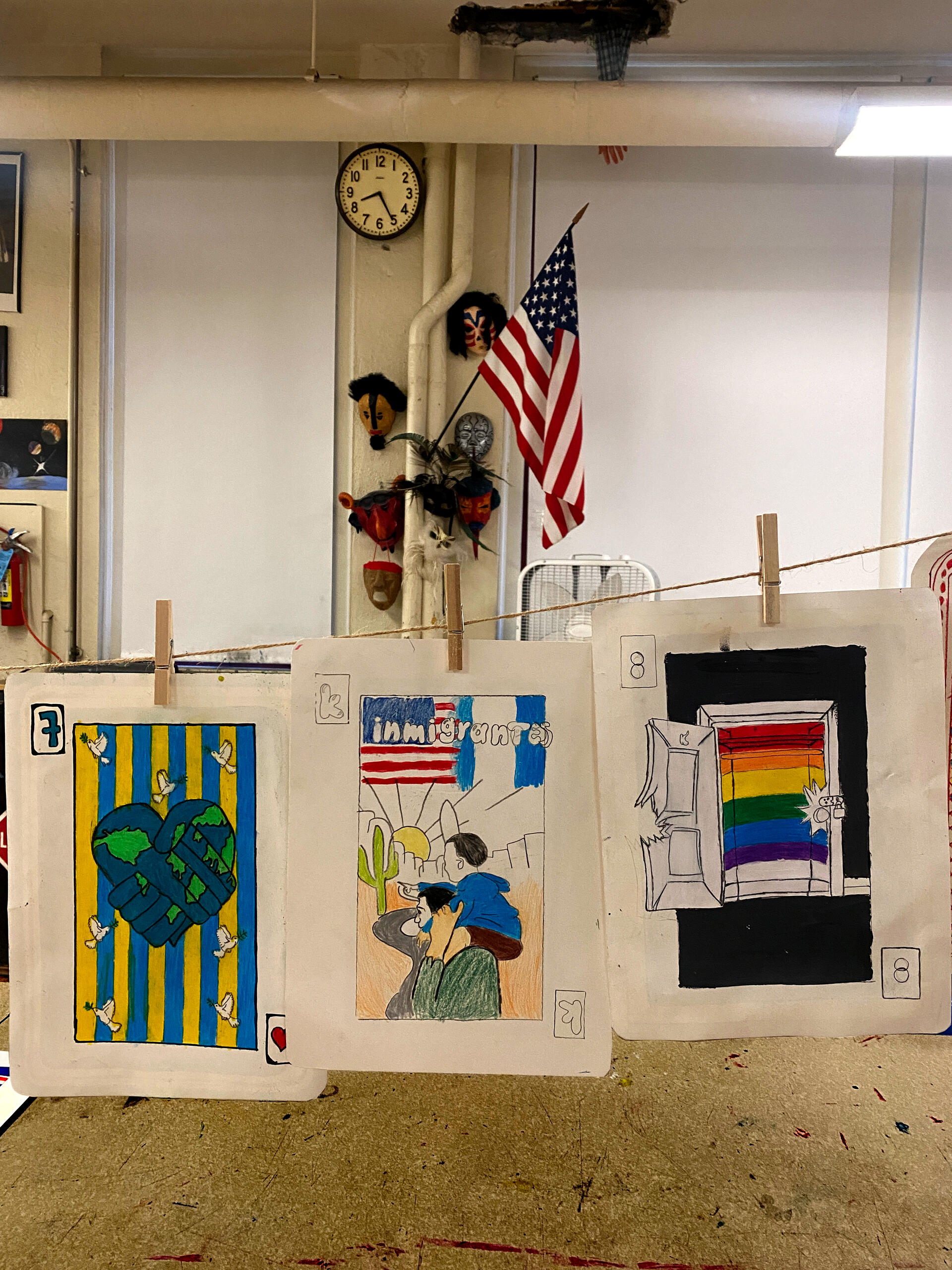 An assortment of drawings in the format of playing cards are suspended in from on a classroom wall with an American flag. Topics of the card include world peace, pro-immigration, and LGBTQ rights.