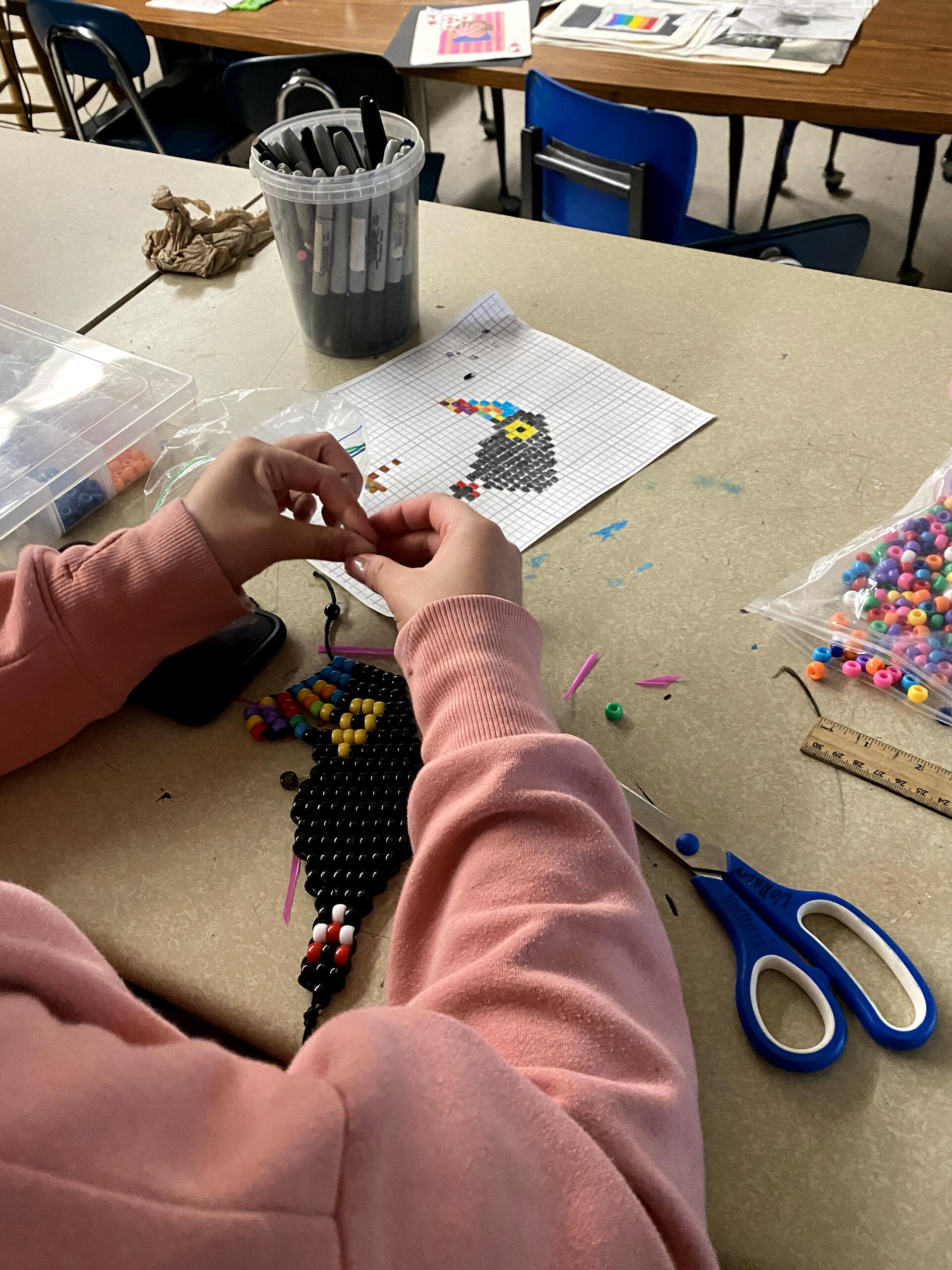 A student's hands are seen working on a pony bead toucan. A drawn design of a toucan on graph paper is seen on the desk as well. Also present are a variety of beads and markers.