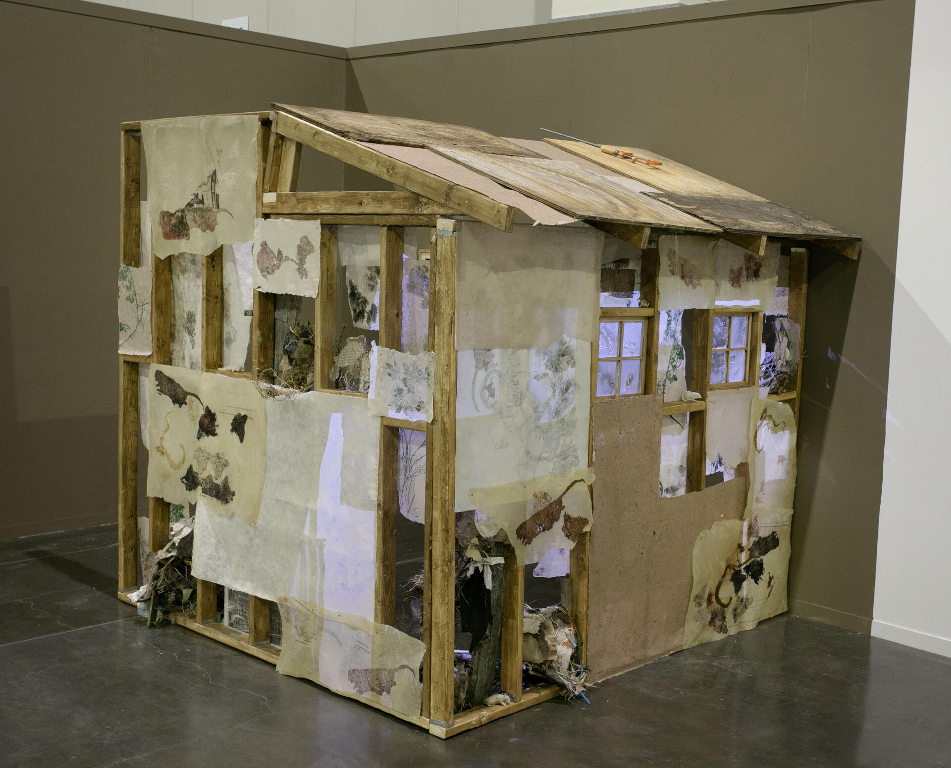 a house-like structue roughly under construction, prints of plants and roadkill line the wall, with a glow comingthrough the windows. The roof is made out of old wood and paper.