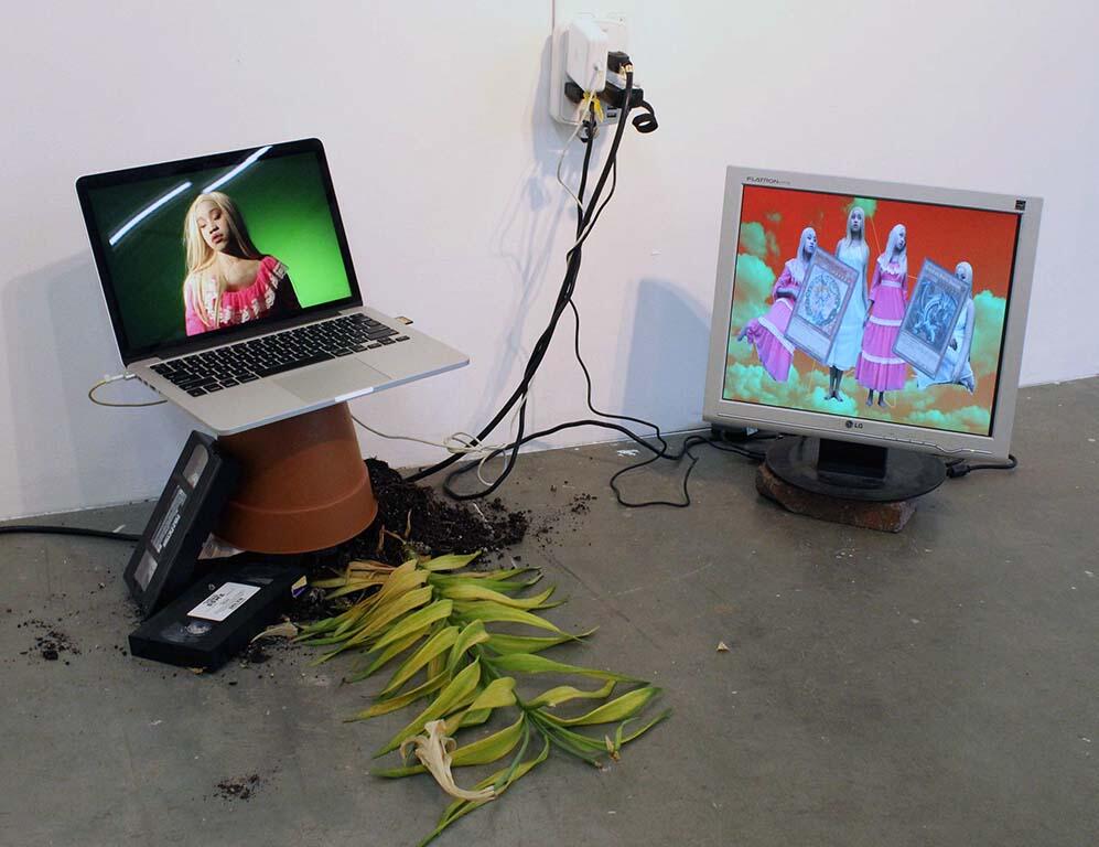 Two screens sit on the floor of the gallery. DARKMAGICIANGIRL appears on a screen to the right and Untitled (Pink Portrait) appears to the left, sitting on top of a terracotta flower pot. There are two VHS tapes to the left of the pot, and a peace-lily with potting soil spilling out from underneath it. 