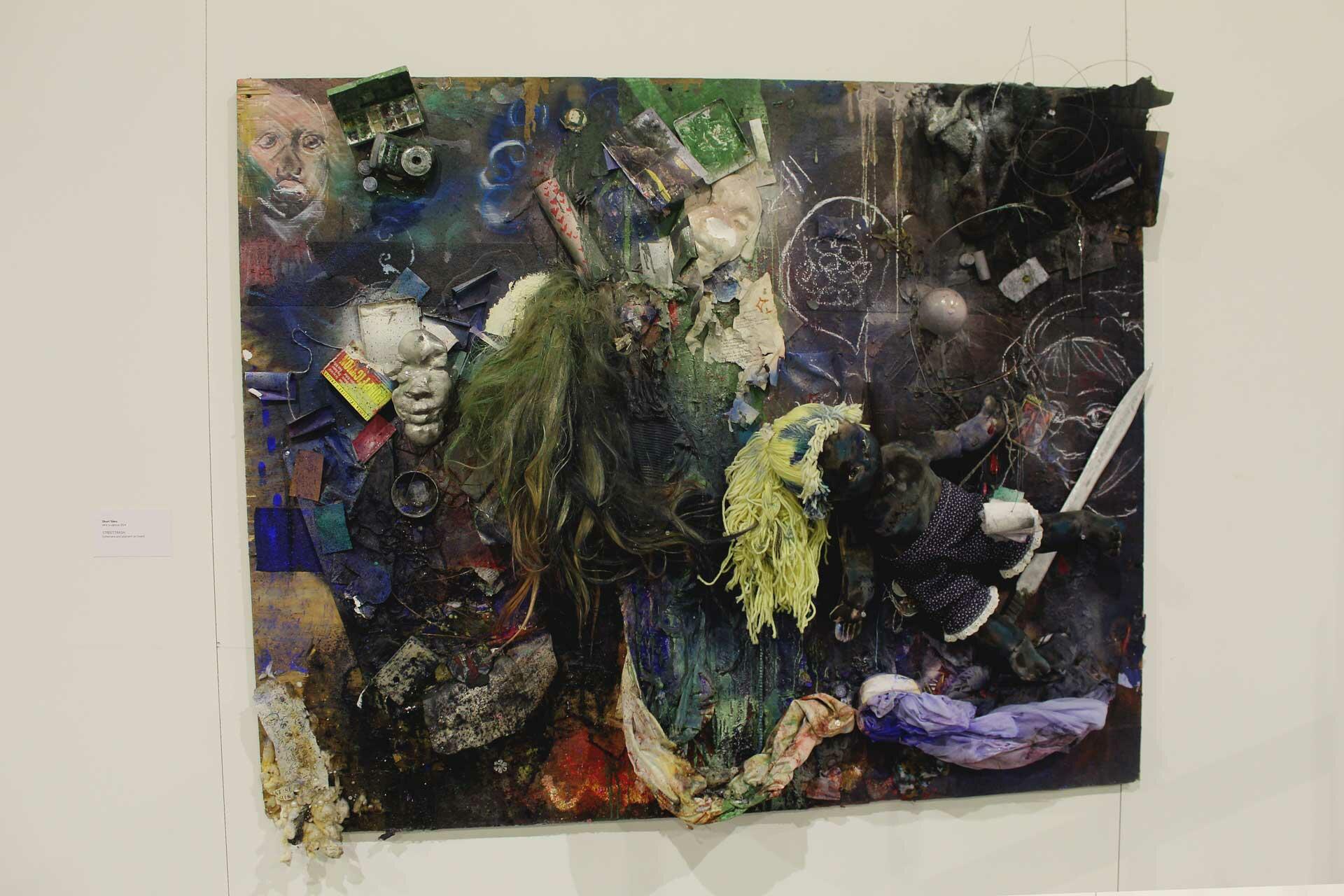 The image depicts a large painting, with dark pigment around the edges and more greenish and blueish shades of pigment towards the middle. To the center-right and upper-left of the painting, there are drawings of faces, and many objects and ephemera attached to the board as well, including a sword, cassette tapes and cases, a small digital camera, a glass orb, a small watercolor compact, and a fabric doll with blond yarn hair. 