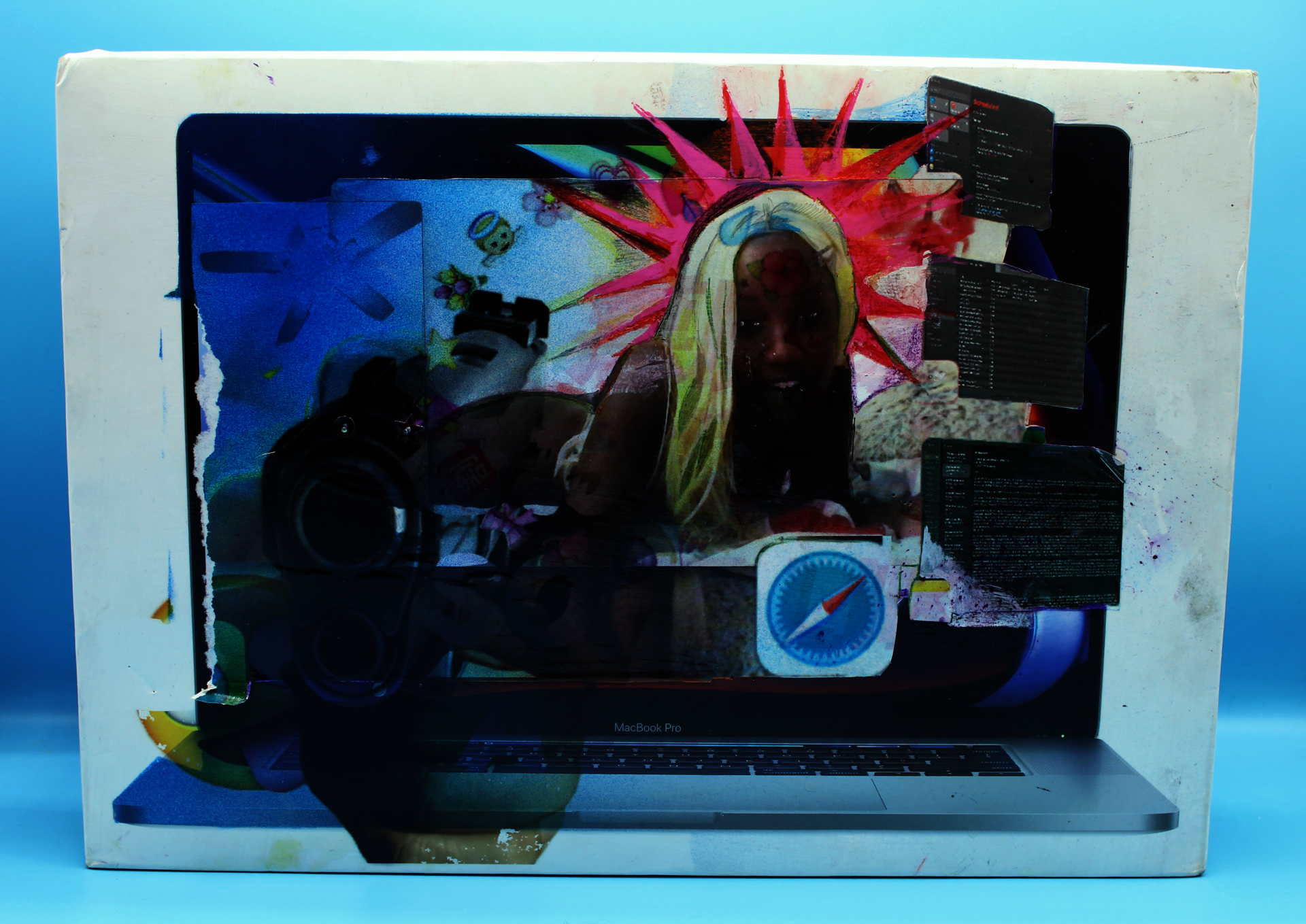 A Macbook Pro 2019 box in a periwinkle light-box. An image of the artist is collaged in the center of the box and surrounded by emojis. There is a hand holding a gun toward the viewer UV printed on the lower-left of the box.