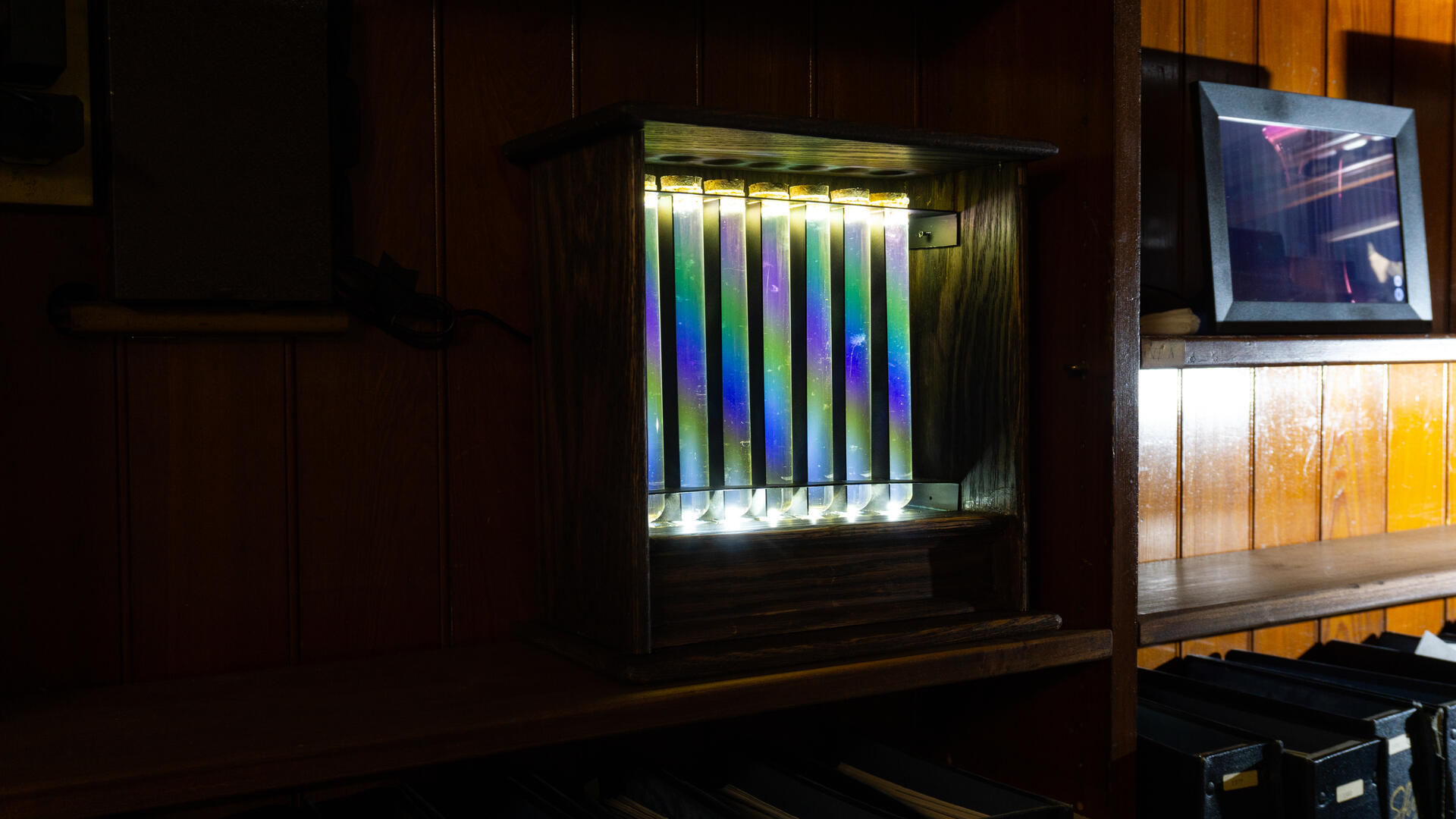 An antique-like wooden box holds a line of test tubes filled with liquids, with iridescent colors forming an arc from the device's arrangement.