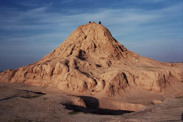 Color photograph of the Assur Ziggurat. What is left of the ziggurat (it is mostly eroded) rises out of sand in a big mound against a blue sky. There are three small figures standing on top.