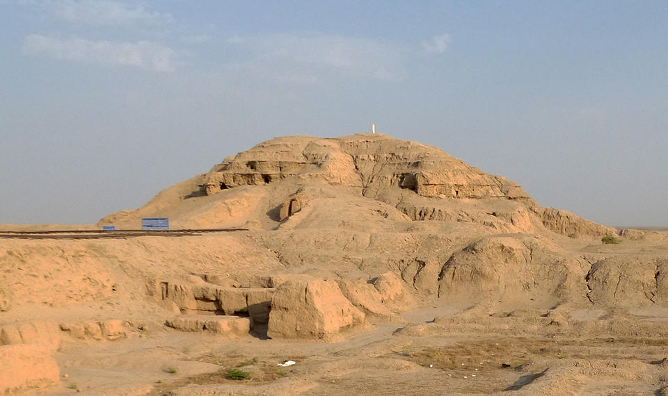 color photograph of the Anu Ziggurat at Uruk. The remains of the stepped pyramid ziggurat emerge from the yellow sand desert, with blue sky above.