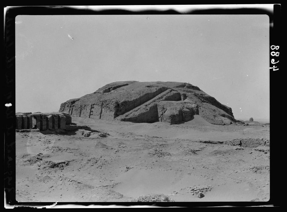 Black and white photograph of the Ziggurat of Ur. The remnants of the ziggurat sit on the horizon line, with sand below and sky above. Only the ziggurat's square base remains, with a triangular staircase.