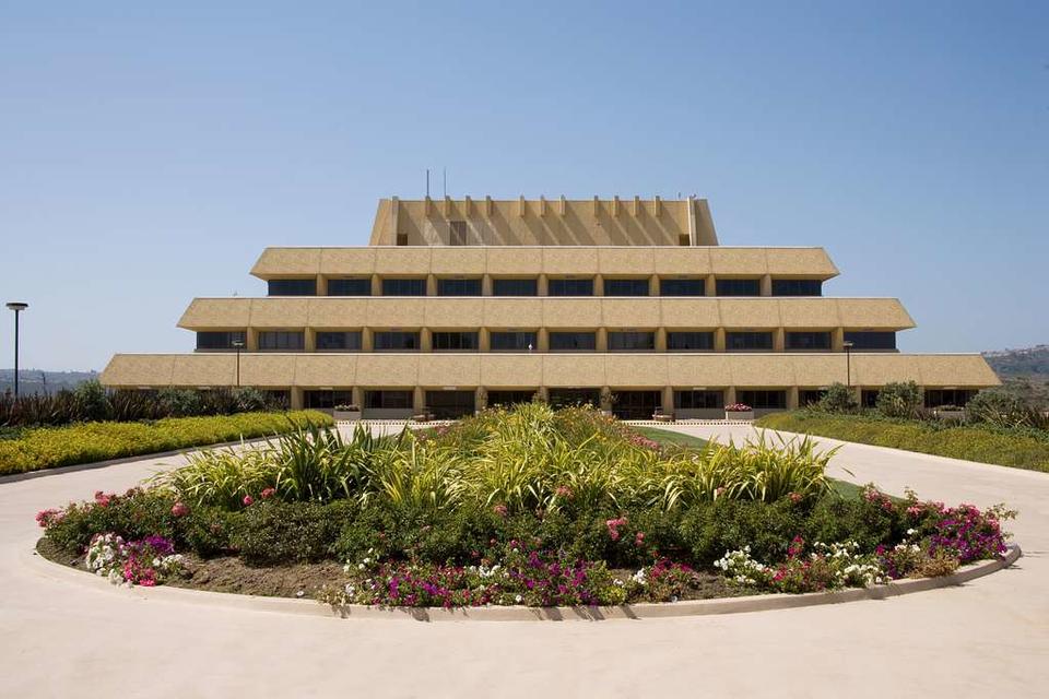 color photograph of the Chet Holifield Federal building in California. Taken from a low angle, there is a lush green planting in the forefront of the composition. In the background, the building, a modernist interpretation of a ziggurat, rises from the horizon line against a blue sky.