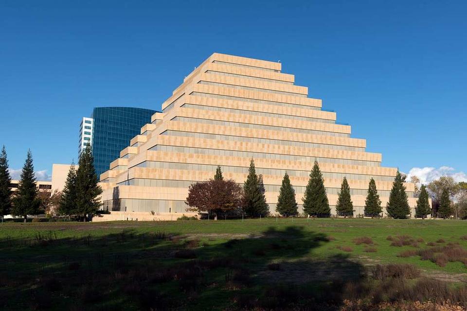 Color photograph of the California Department of General Services building, a 10-story contemporary ziggurat building. Taken from a 3/4 view, this photograph shows of the stepped sides of the tan building against a blue sky.