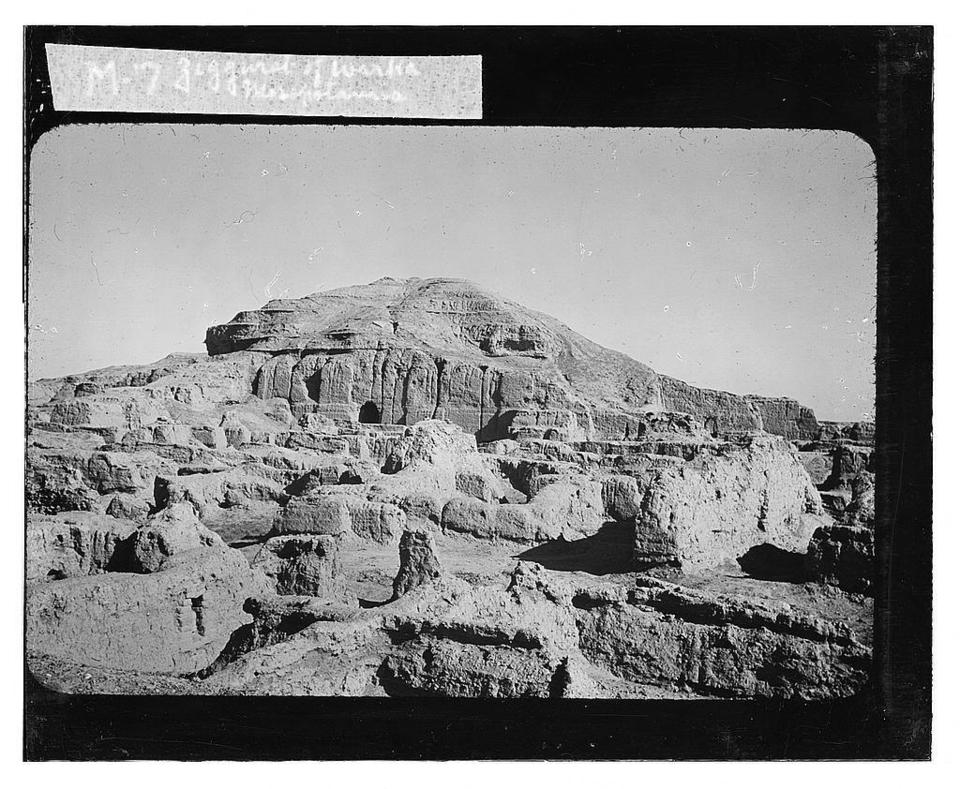 black and white photograph of the Ziggurat of Warka. Foreground is scattered stone blocks in a desert landscape. Beyond is the ruins of the Warka Ziggurat.