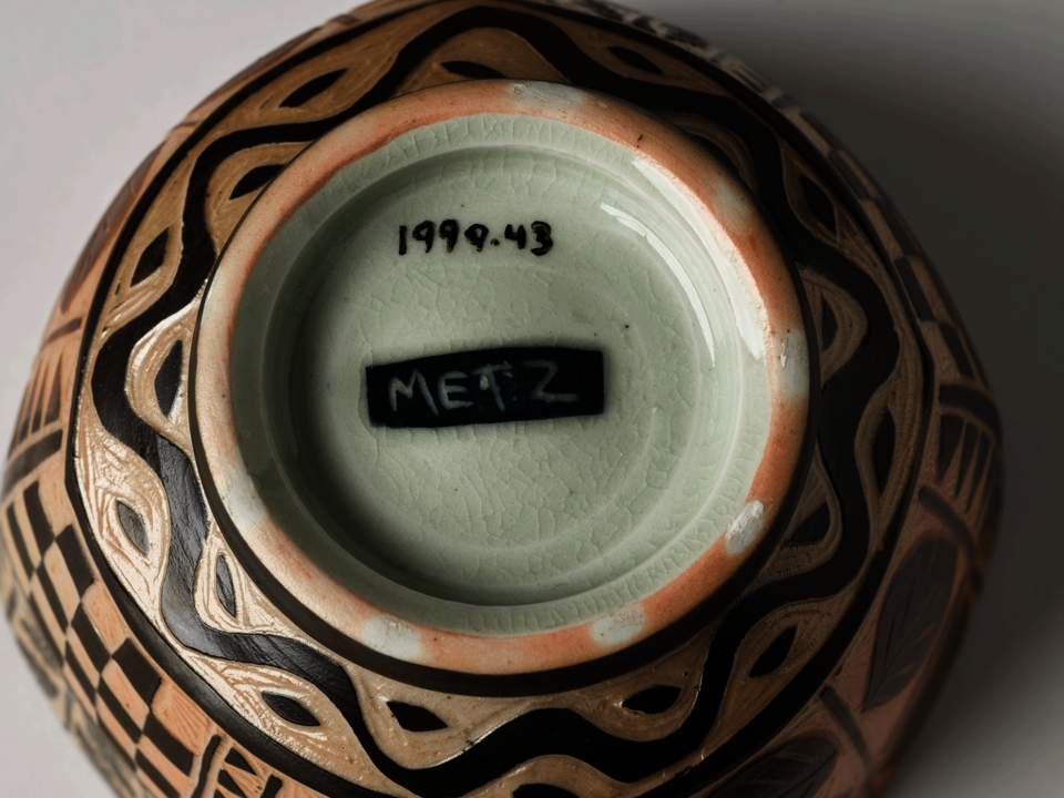 Underside of bowl. Wavy line and leaf-shaped patterning scratched into clay, circling the foot of the bowl. Light green interior of the foot has a black rectangle in the center with the name “Metz” painted inside. Accession number is visible.