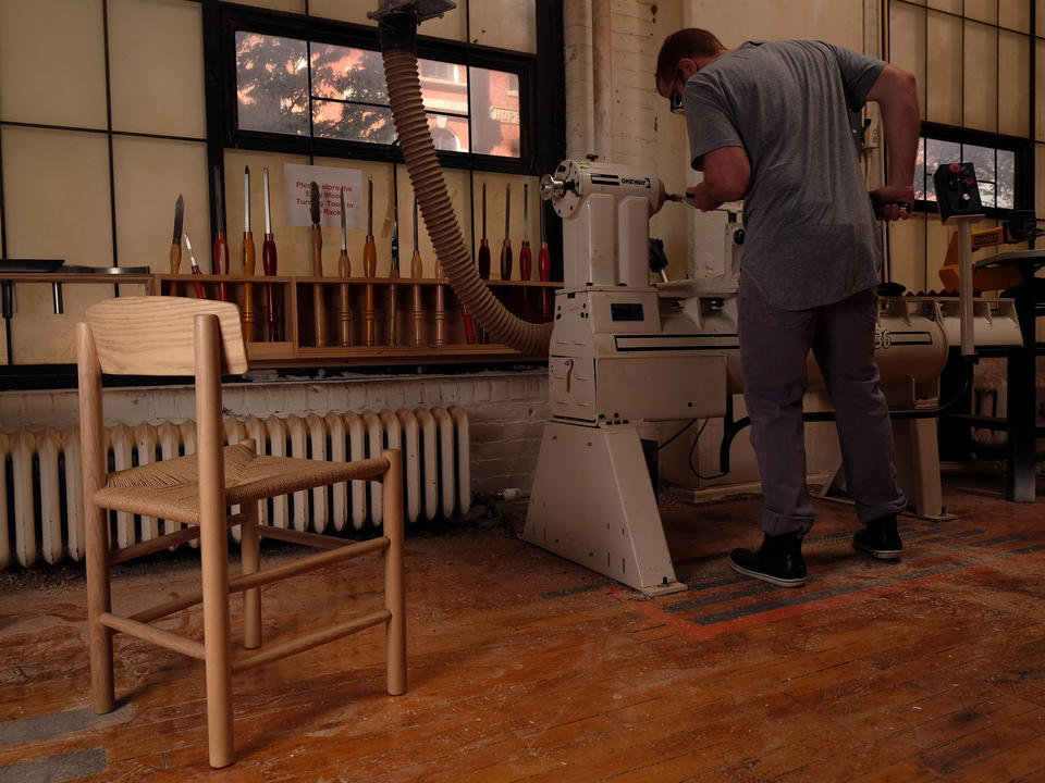 An image of Analog |Digital | Analog, a replica of the J39 chair, seated in front of me turning parts for a new one at the wood lathe in the Furniture Design wood shop in the Metcalf building.