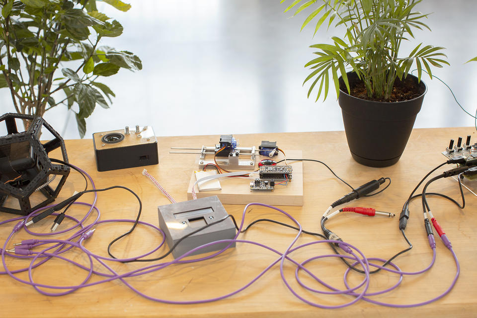A close view of several electronic instruments and a plant arranged on a light wooden tabletop. Audio cables and wires connect them and spiral around.