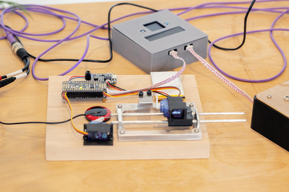 A close view of a robotic music box. A small motorized plastic lever is angled upward facing the music box tines. In the background, a small gray box can be seen. Wires connect the box and a circuitboard on the robotic music box.