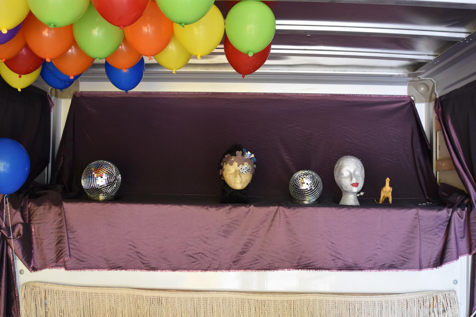 Interior of a U-haul, draped in shiny purple fabric, with two disco balls and two foam mannequin heads and a toy dinosaur sitting on a ledge. Balloons hover on the ceiling.