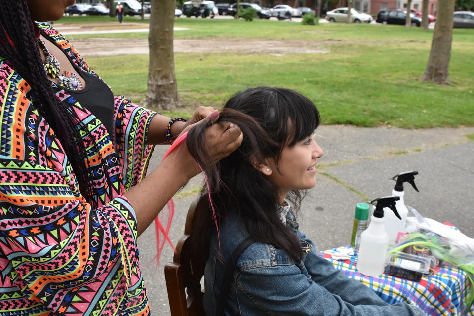 Female teen sits as another female teen in a bright shirt and necklace braids a bright pink hair extension into her hair. They are outside in a park.