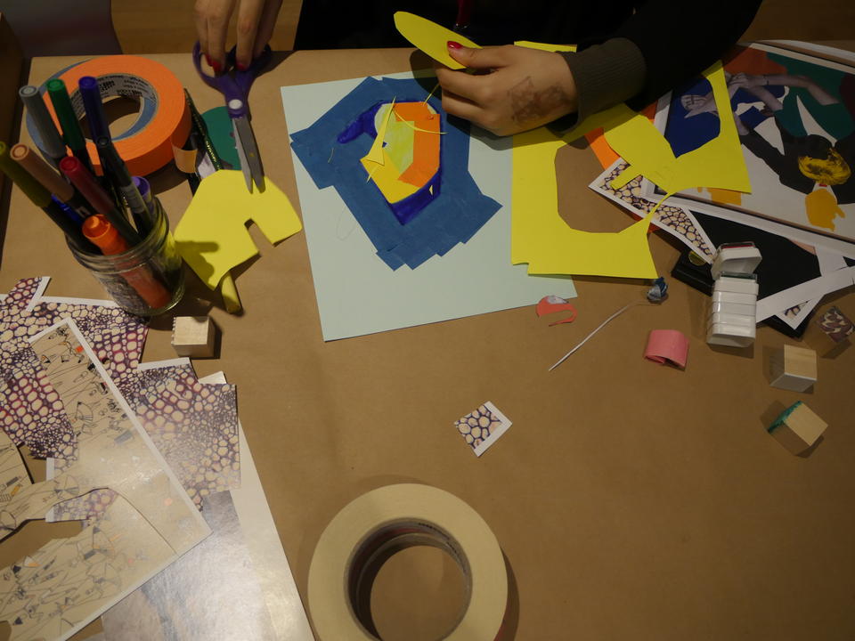 overhead color photo of a table covered in brown paper, strewn with colorful art supplies; a pair of hands grasp a yellow paper circle and a pair of scissors in the top of the image