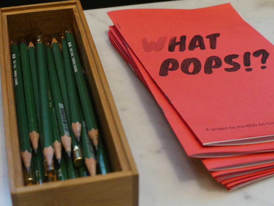 close-up color photograph of a box of green pencils and pile of salmon colored booklets that say 'What Pops!?'