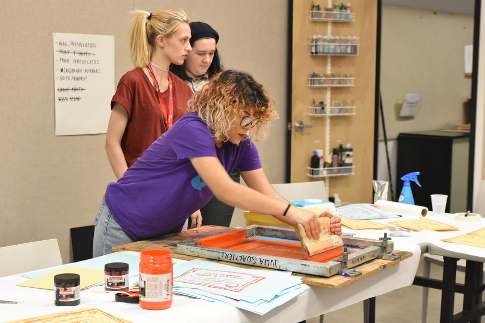 Three teens screenprinting paper with bright orange ink.