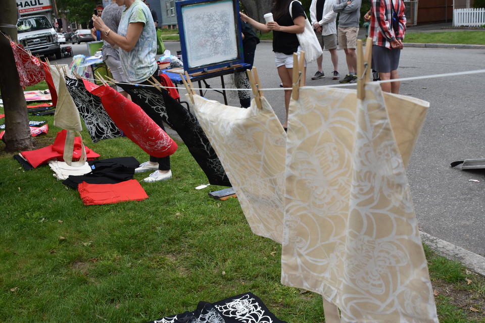 a clothesline of canvas totebags and red and black handkerchiefs hand to dry after being screen printed on. People stand in the background, near a screen printing screen. 