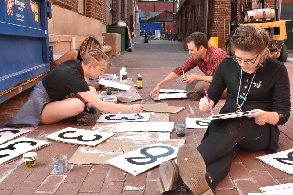 Three teens and one adult male sit on the ground in an alleyway, painting large black numbers onto pieces of cardboard.