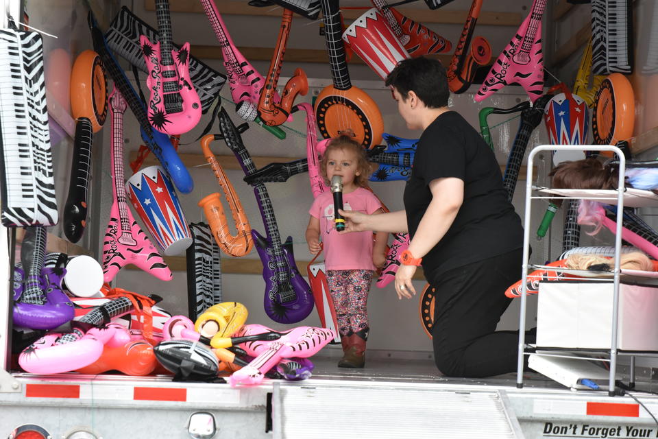 An adult holds a microphone up to a small child in a pink shirt and cowboy boots. They are in the back of a U-haul that is filled with colorful inflatable rock instruments.