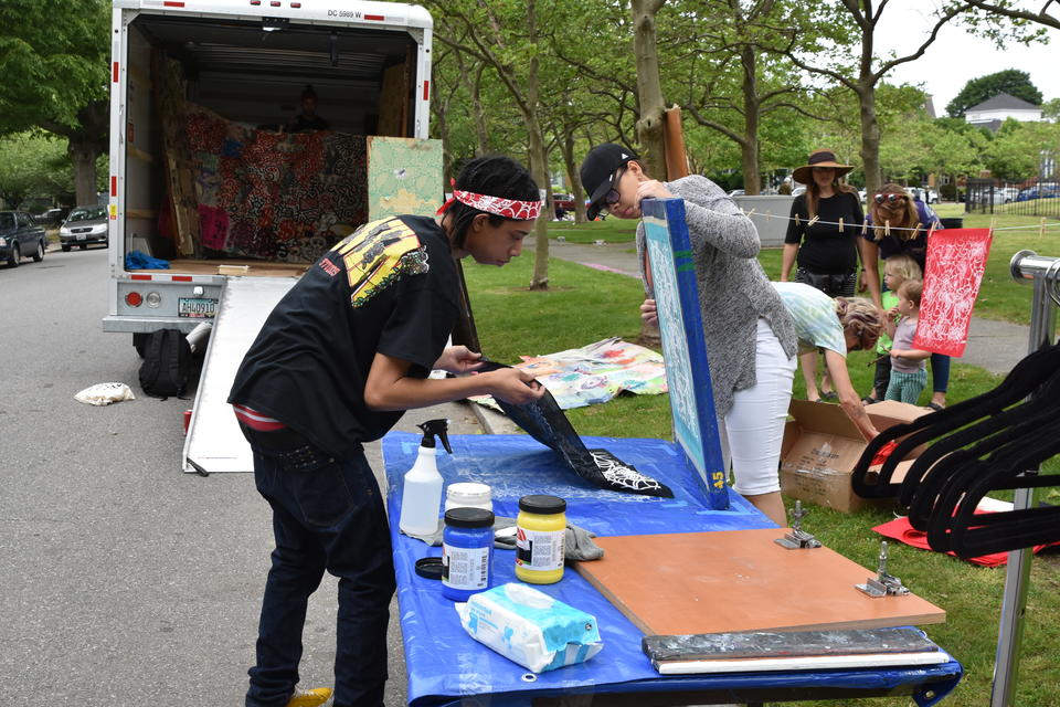 Two black teens screen print a black bandana. They are outside by a park and an open U-haul truck with artwork inside.