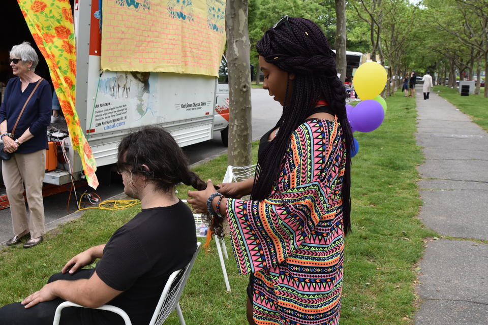 A black teen with long braids and a colorful shirt braids the hair of a seated teen. They are outside in a park near a U-haul with a large mural on it.